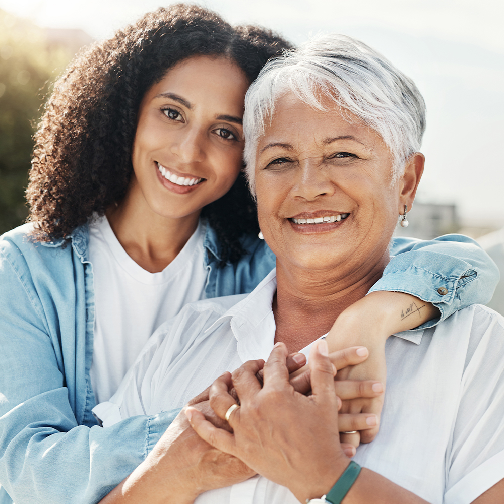 Older woman with short gray hair smiling and being embraced from behind by a younger woman, both outdoors with a blurred crowd in the background.