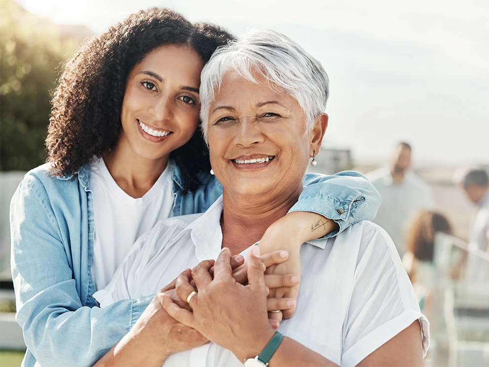 Older woman with short gray hair smiling and being embraced from behind by a younger woman, both outdoors with a blurred crowd in the background.