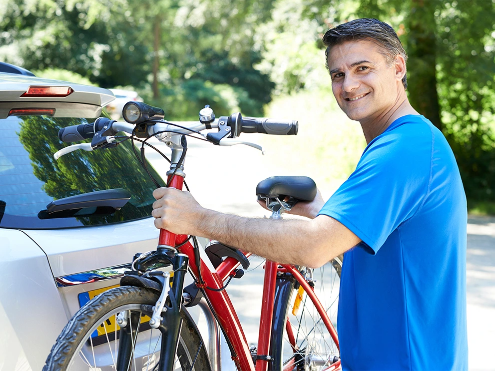 Man in a blue shirt smiling while securing a red bicycle to a rear-mounted rack on a car, with a wooded area in the background.