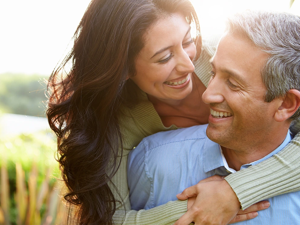 Smiling woman embracing a smiling man from behind in an outdoor setting with sunlight and greenery in the background.