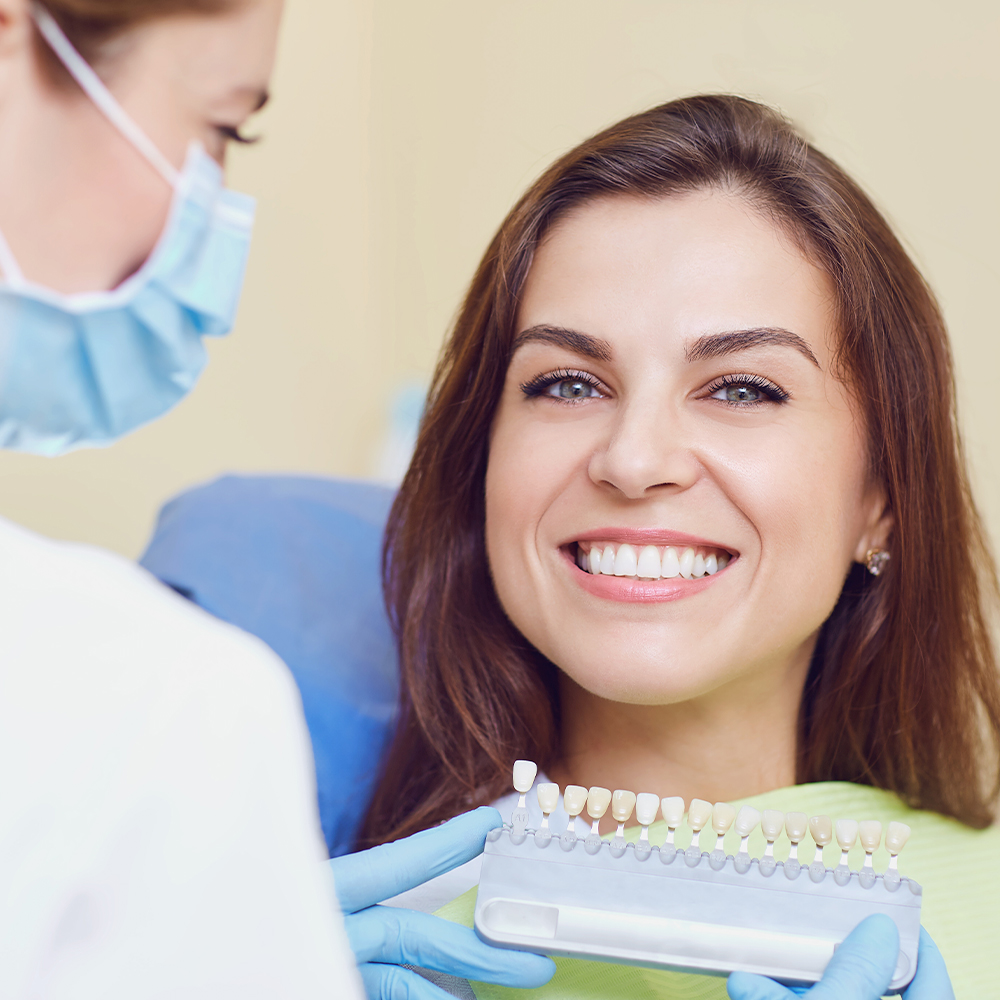 Female dental patient smiling in a dental chair while a gloved dentist holds a tooth shade guide next to her teeth for color matching, commonly used in cosmetic dentistry.