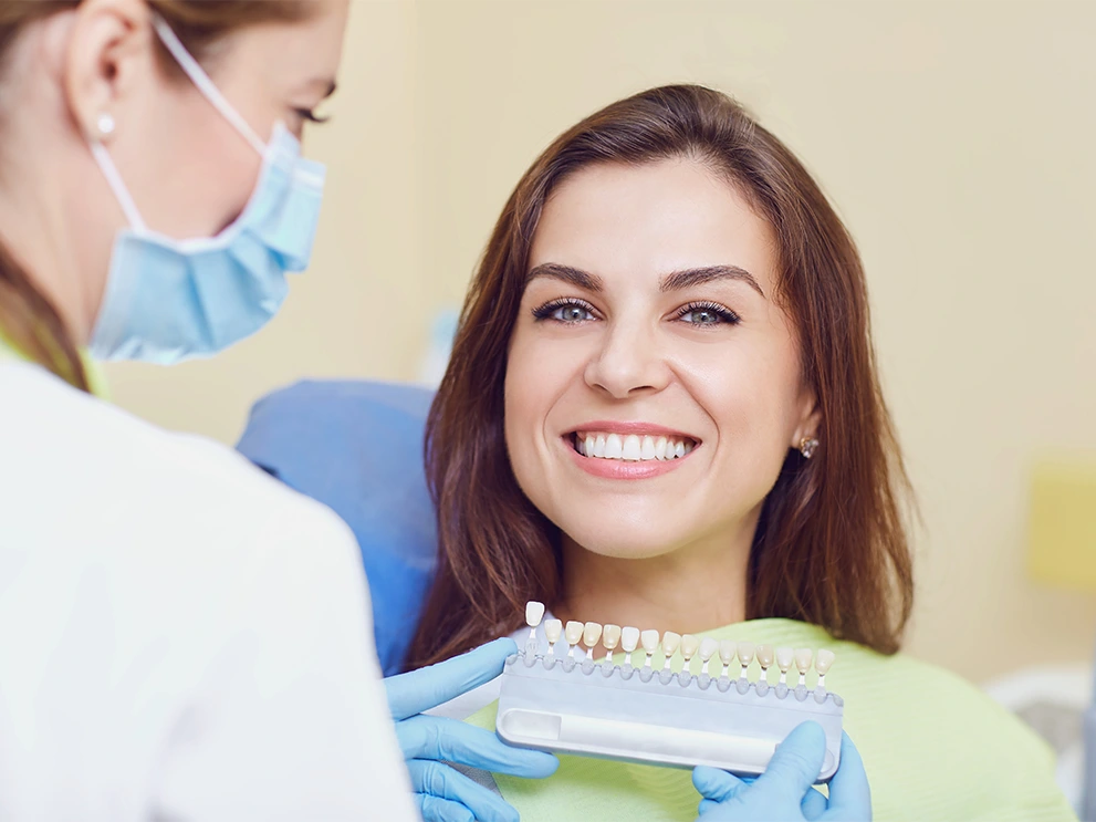 Female dental patient smiling in a dental chair while a gloved dentist holds a tooth shade guide next to her teeth for color matching, commonly used in cosmetic dentistry.