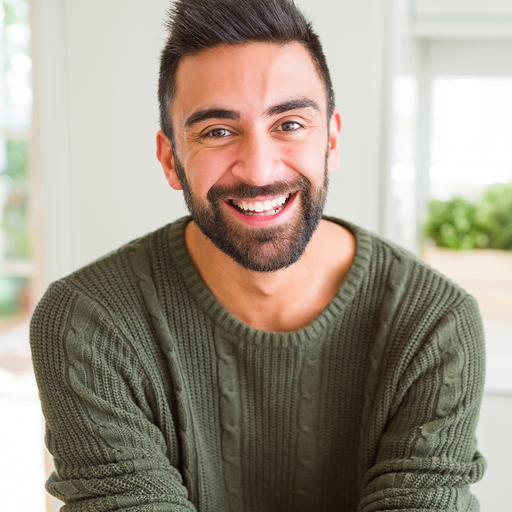 Bearded man wearing a green knitted sweater smiling warmly indoors, with a blurred background of potted plants and natural light.