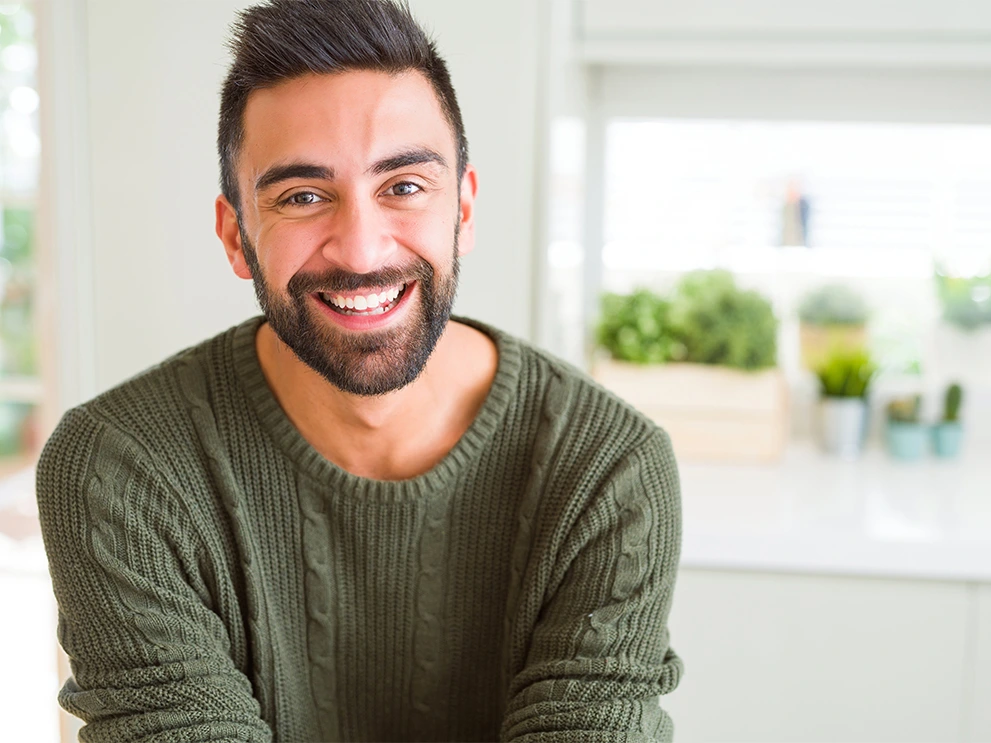 Bearded man wearing a green knitted sweater smiling warmly indoors, with a blurred background of potted plants and natural light.